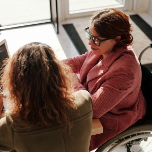 Two women working on laptops, one using a wheelchair, collaborating in a modern café.